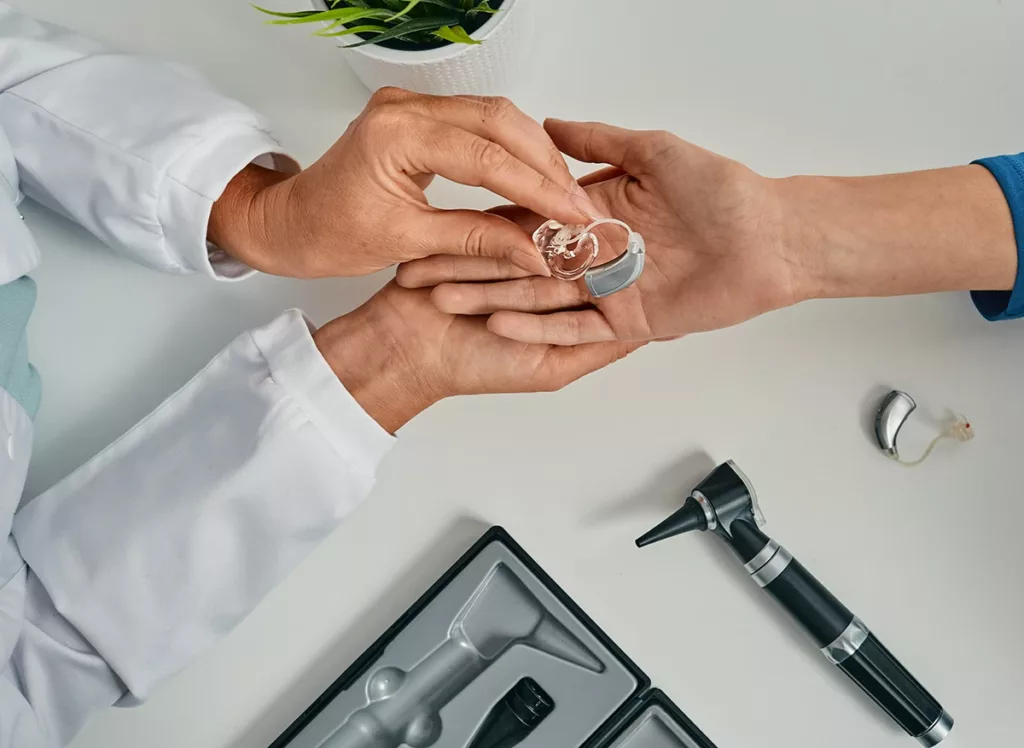 Top view of hearing health care specialists putting modern BTE hearing aid in patient hand for treatment hearing loss at hearing clinic.